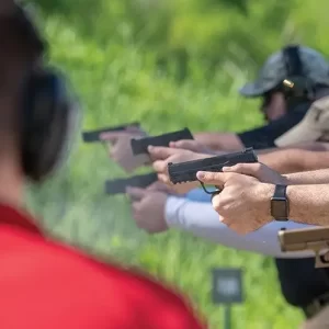 Photo of a Range Safety Office in a Red Shirt watching over a class of students holding pistol, aiming down range. There is 5 students and 1 instructor.