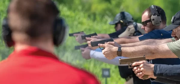 Photo of a Range Safety Office in a Red Shirt watching over a class of students holding pistol, aiming down range. There is 5 students and 1 instructor.