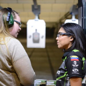 Two Female shooters standing in a shooter's booth at the range. One of them is a firearms instructor the other is a shooter student. The instructor is in a black shirt with black safety glasses. The student is in a tan shirt with green ear protection and black eye protection. There is a black silhouette target down range.