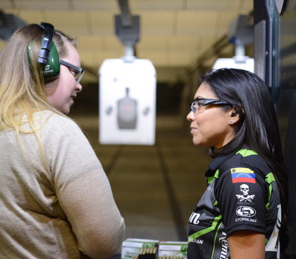 Two Female shooters standing in a shooter's booth at the range. One of them is a firearms instructor the other is a shooter student. The instructor is in a black shirt with black safety glasses. The student is in a tan shirt with green ear protection and black eye protection. There is a black silhouette target down range.